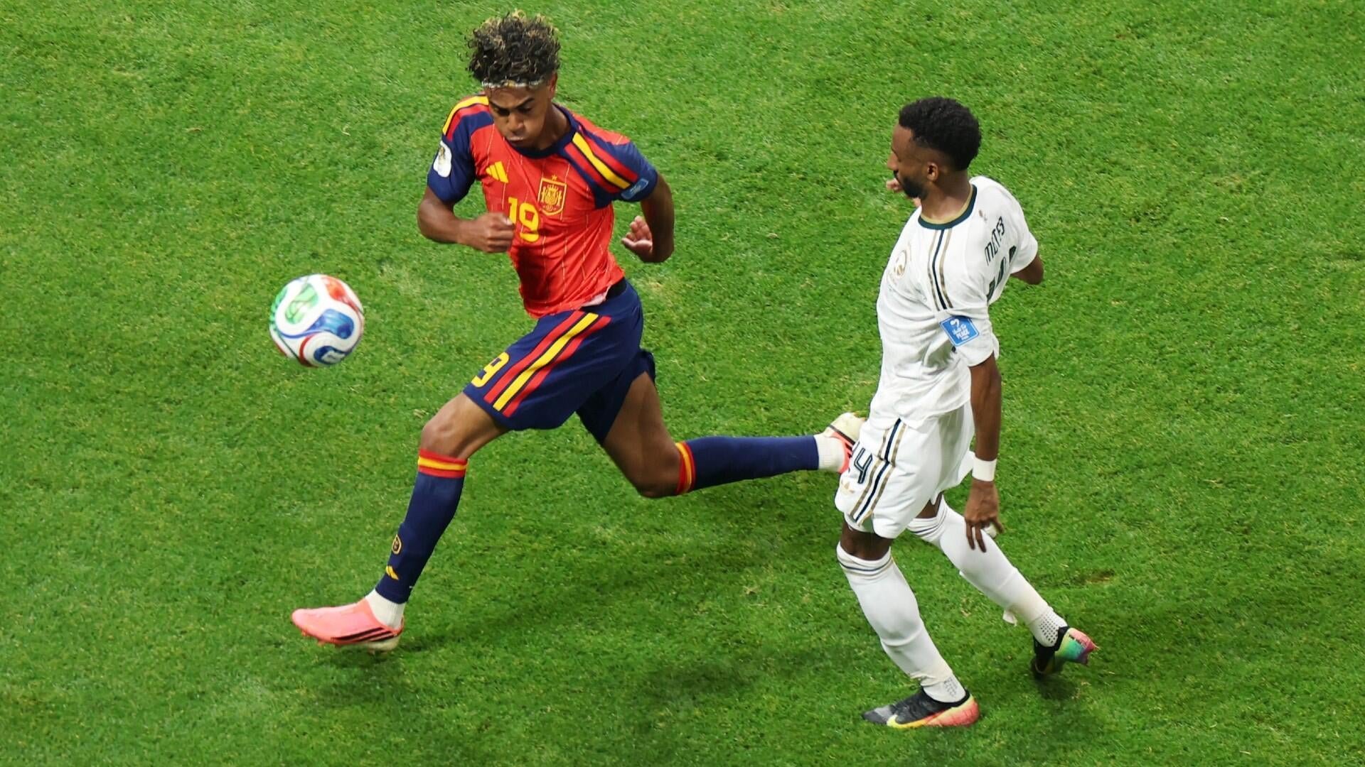 A soccer player in a red Spain jersey (number 19) chases a ball across green grass while being pursued by an opponent in a white jersey.