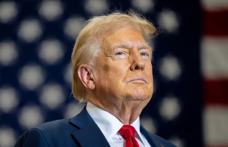 A man with blonde hair in a dark suit and red tie speaks at a microphone, with an American flag visible in the background.