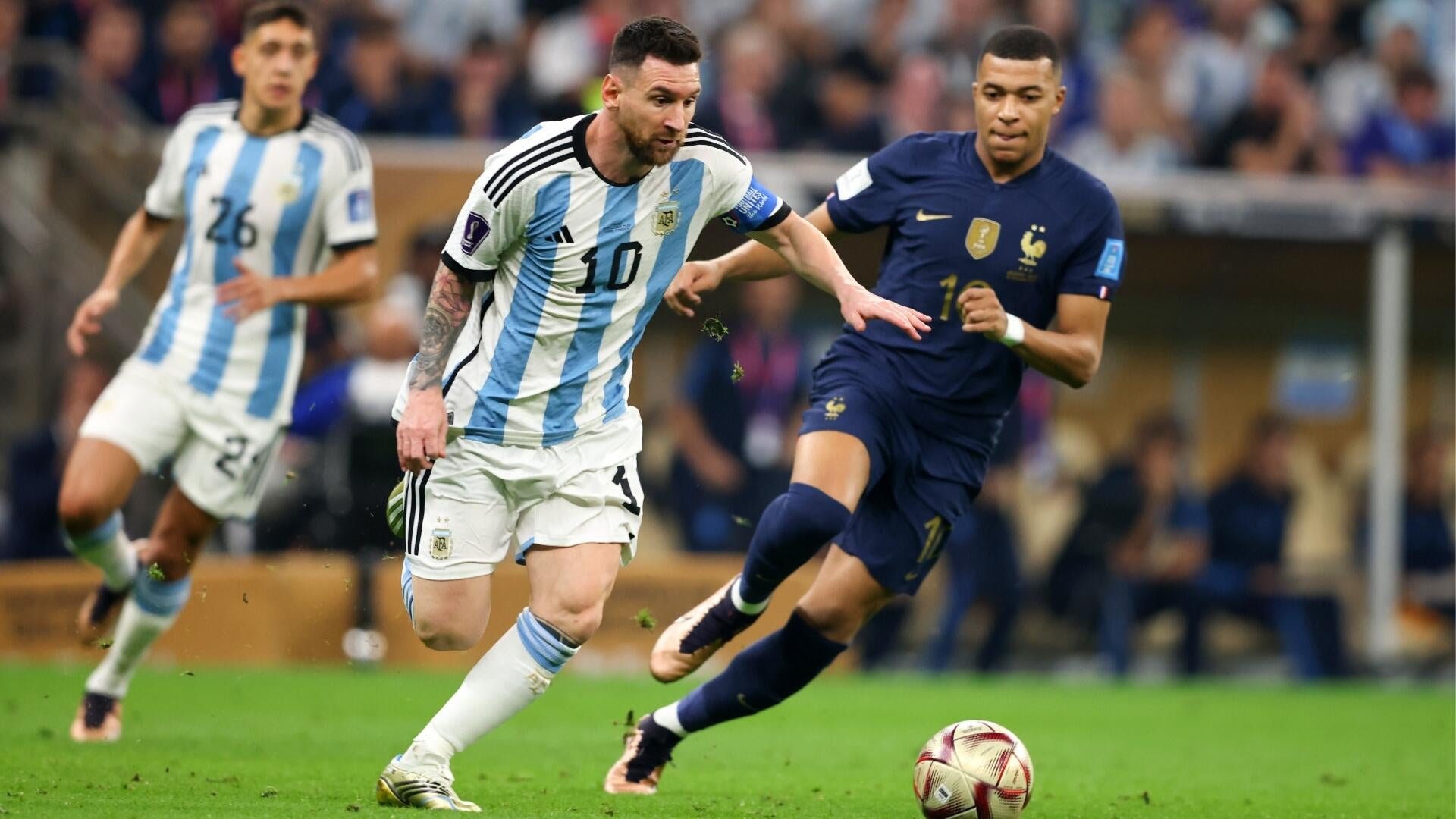 Two soccer players in light blue and white striped jerseys compete against an opponent in a dark blue jersey for the ball on a grass pitch.
