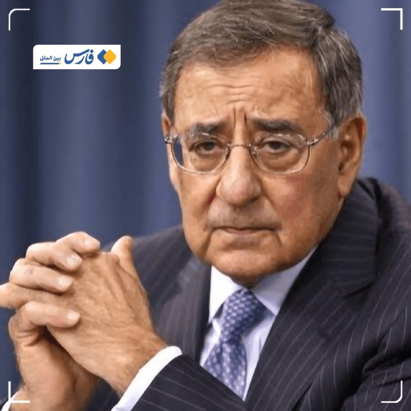A man with gray hair and glasses, wearing a dark pinstripe suit and blue tie, sits with his hands clasped in front of a blue curtain backdrop.