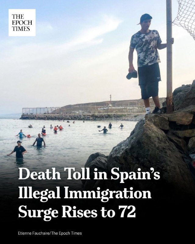 A man stands on rocky shore near a pole while numerous people wade through ocean water toward land, with a headlined news graphic reading "Death Toll in Spain's Illegal Immigration Surge Rises to 72."