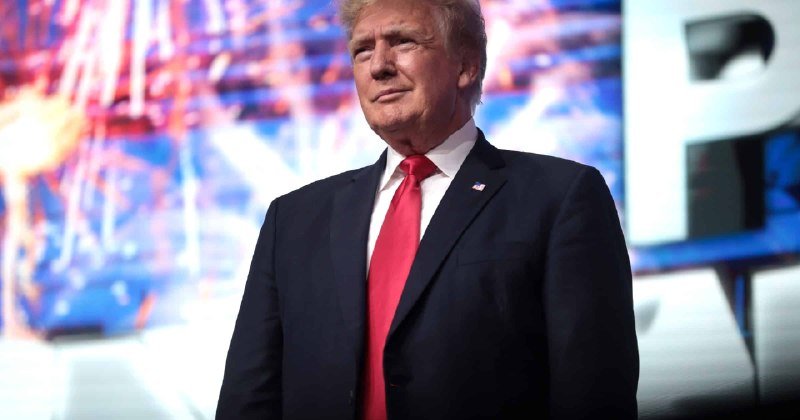 A man in a dark suit and red tie stands on a stage with a U.S. flag pin, smiling toward the audience against a blue patriotic backdrop.