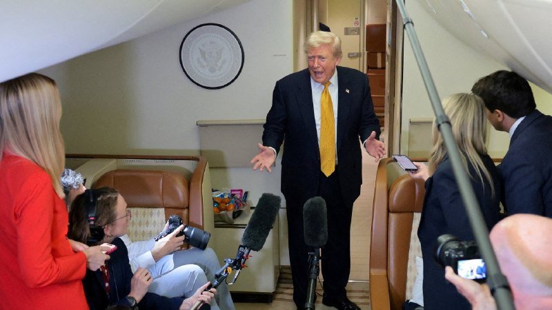 A man in a dark suit and yellow tie gestures with open arms toward reporters holding microphones and cameras aboard an aircraft featuring the presidential seal on the wall.