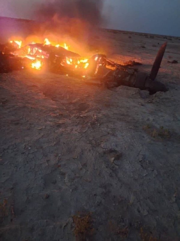A helicopter wreckage burns with visible flames and thick black smoke on a barren dirt field.