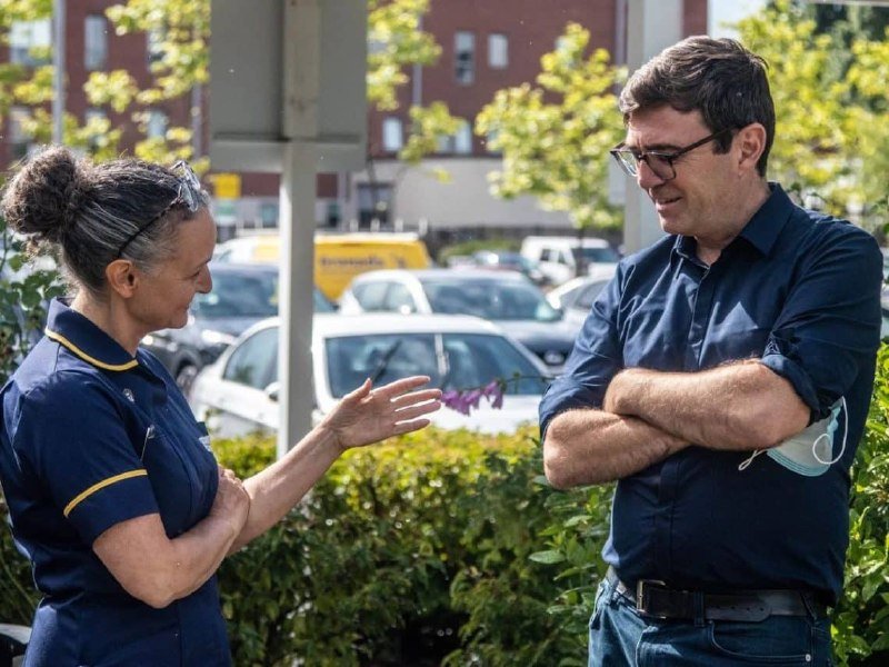 A woman in a dark uniform gestures while speaking to a man in a navy shirt and glasses, who stands with arms crossed outdoors near parked cars.