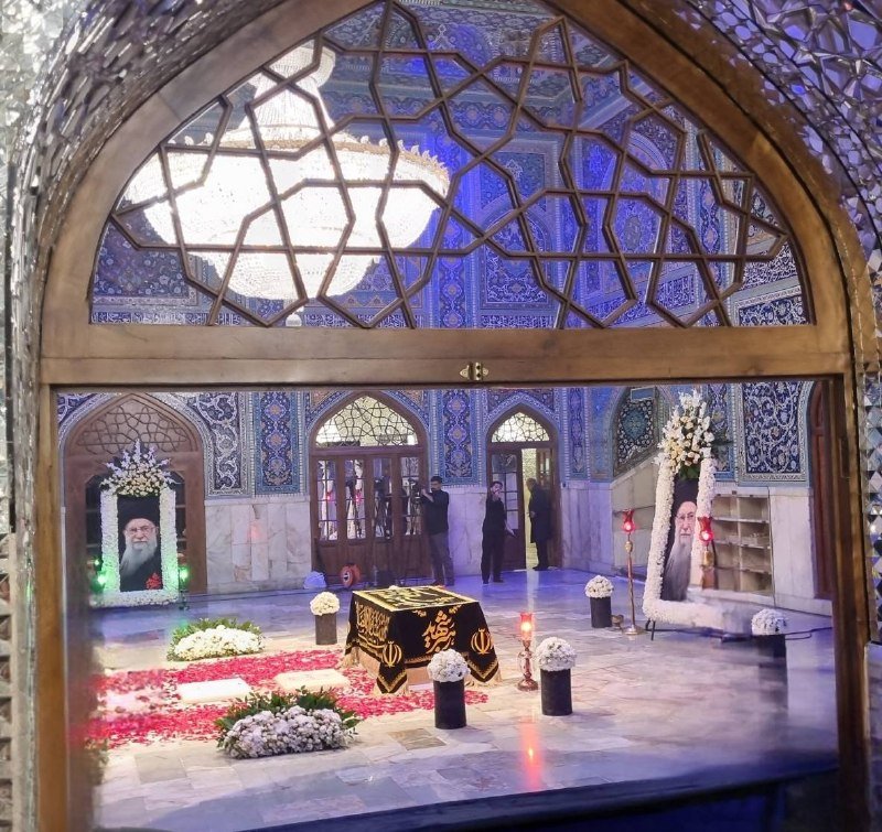 Viewed through an arched stained-glass window, an ornate tiled shrine interior features a draped tomb, framed portraits of bearded men surrounded by flowers, and scattered visitors.