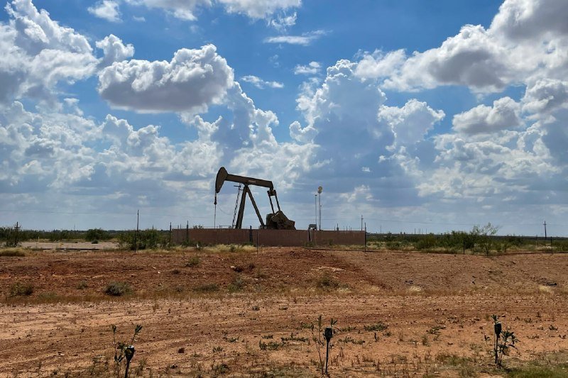 An oil pumpjack operates in a fenced dirt lot under a bright blue sky filled with white clouds.