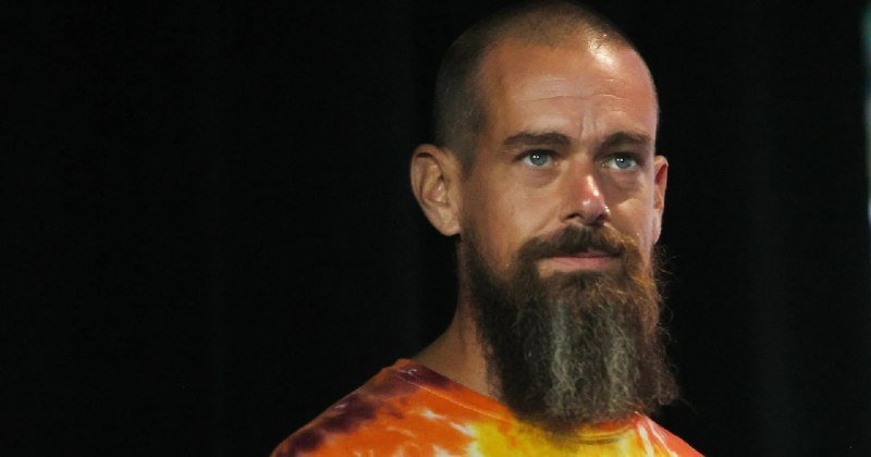 A bald, bearded man wearing an orange and yellow tie-dye shirt looks toward the camera against a dark background.