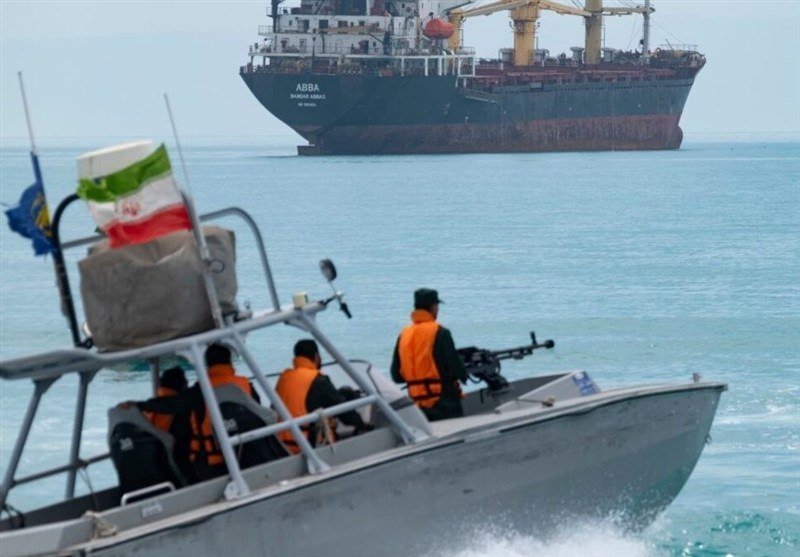 Armed personnel in orange life vests ride a small patrol boat displaying the Iranian flag, with a large tanker ship visible in the background on open water.