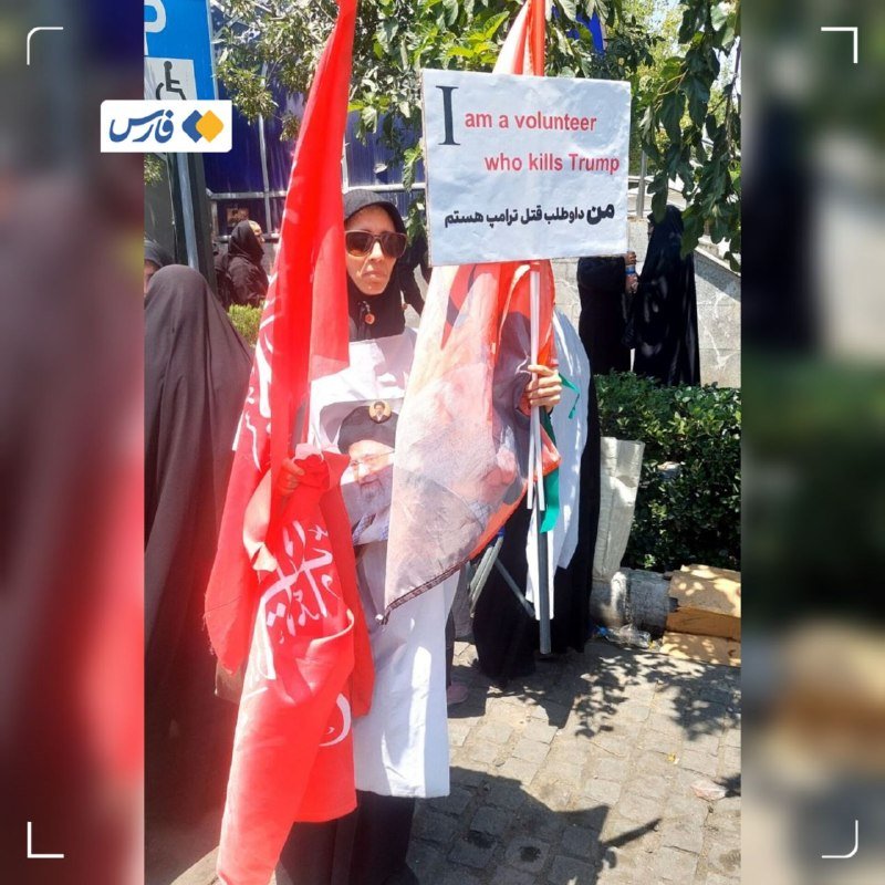 A woman in a black chador and sunglasses holds red flags and a sign reading "I am a volunteer who kills Trump" at an outdoor gathering.
