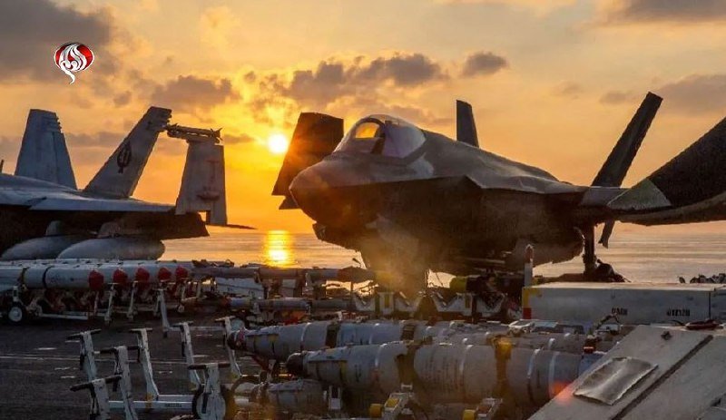 Military fighter jets and rows of mounted munitions sit on a carrier or airbase flight deck at sunset.