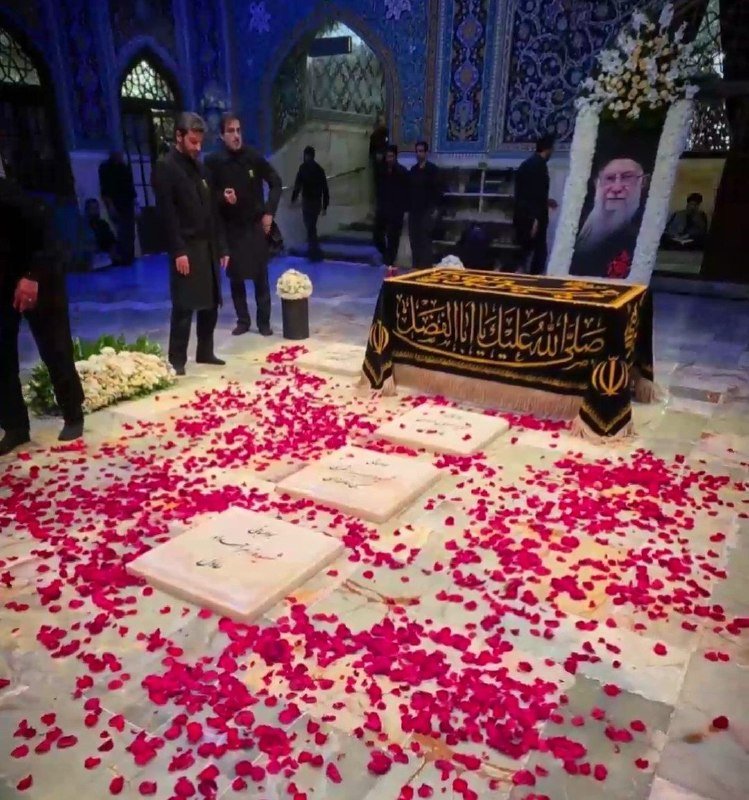 Men in black stand beside an ornately decorated shrine containing a black and gold draped structure and a framed portrait, with red rose petals scattered across the floor.