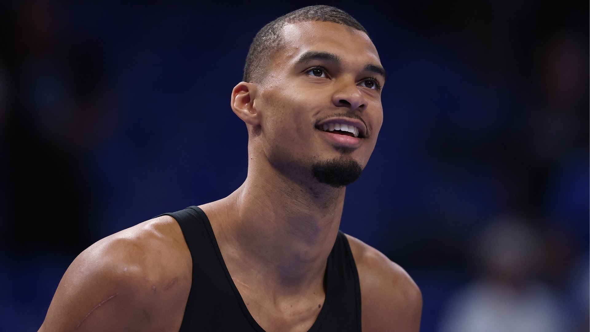 A young man in a black tank top smiles while looking upward against a blurred dark blue background.