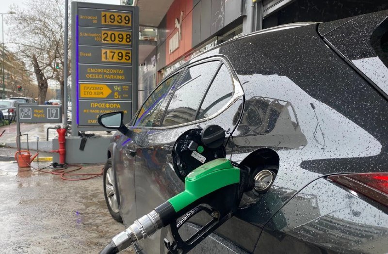 A dark car is refueling with a green-and-black nozzle at a station displaying fuel prices in Greek, including unleaded at 1.939 and diesel at 1.795.