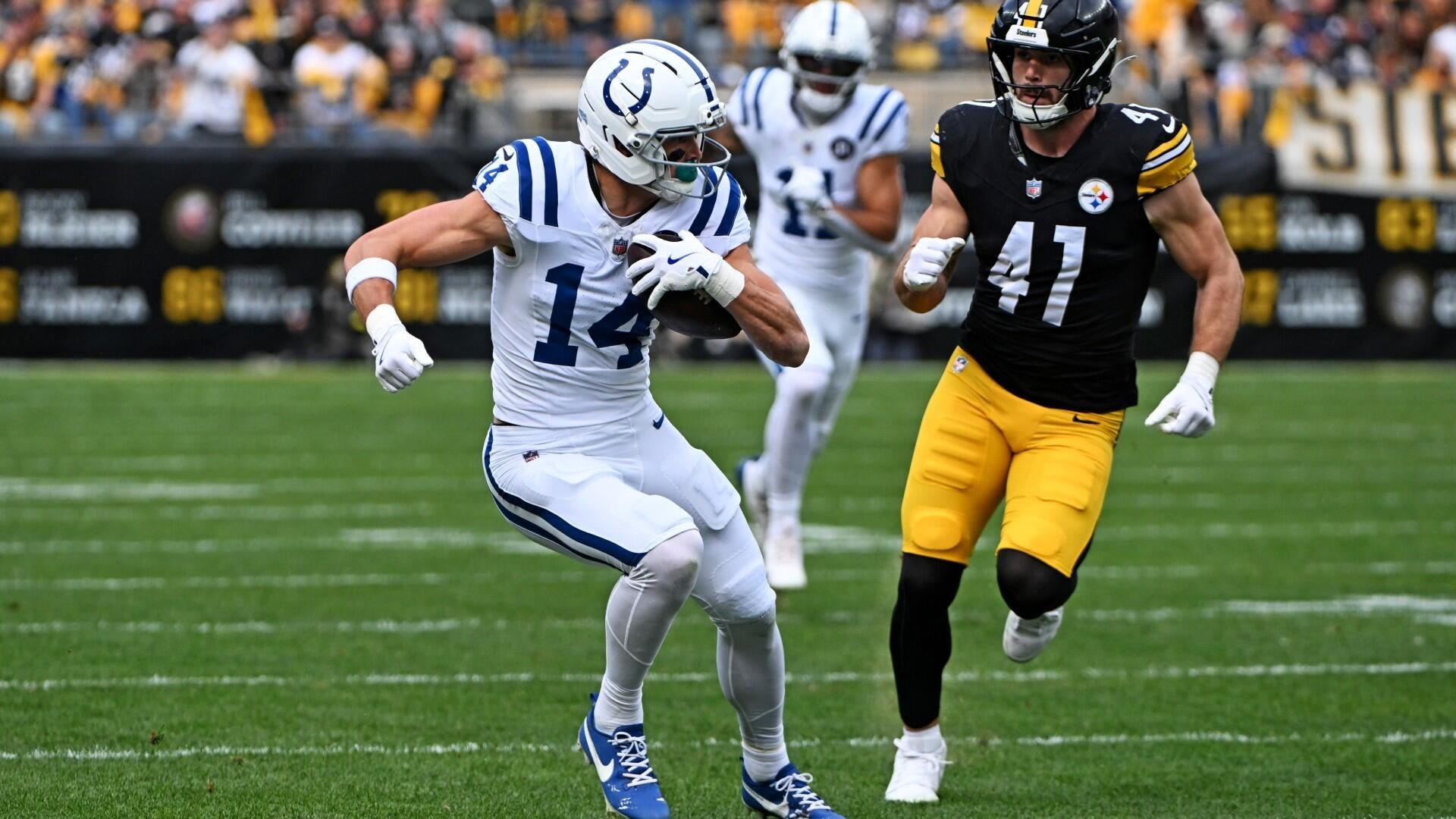 A football player in a white uniform carrying the ball runs downfield while being pursued by a defender in a black and yellow uniform.