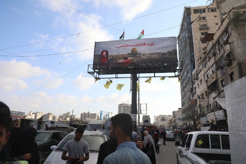 A large billboard displaying Arabic text, flags, and a domed structure hangs over a crowded street filled with pedestrians and vehicles, with weathered buildings lining both sides.