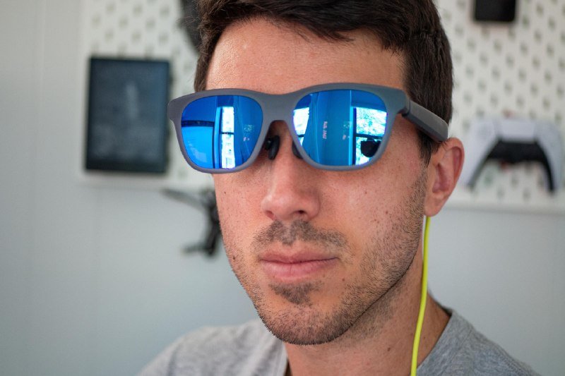 Close-up of a man wearing blue mirrored sunglasses and yellow earphones, with a white game controller visible on a shelf behind him.