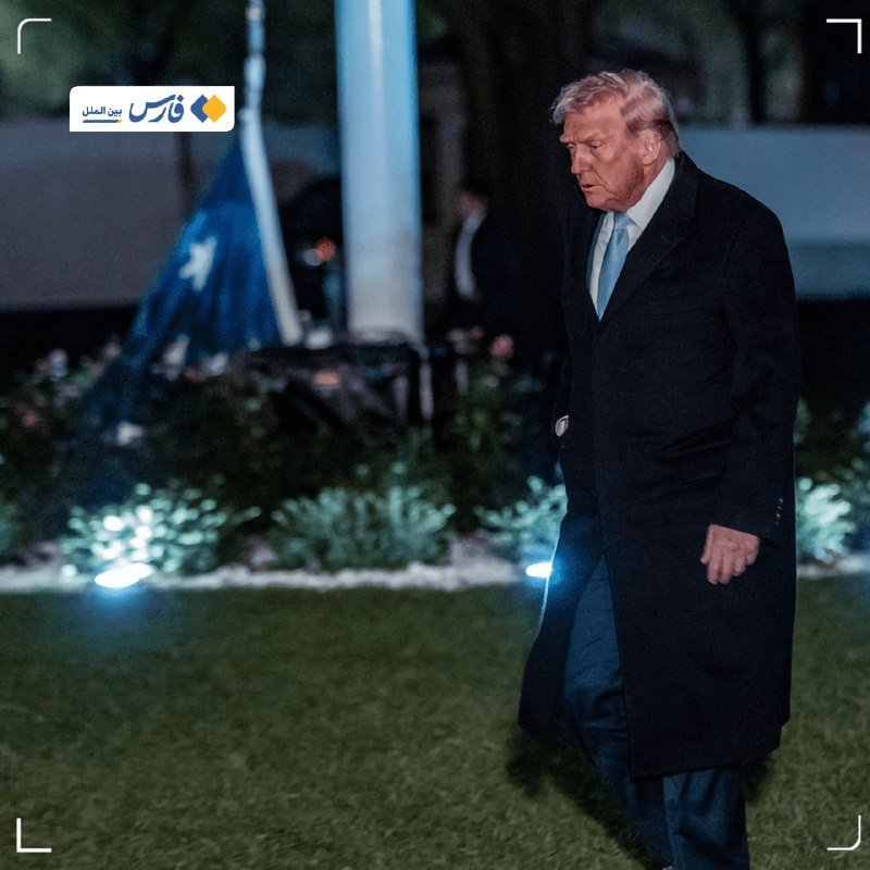 A man in a dark overcoat and blue tie walks across a lawn at night, looking downward, with a partially visible flag and illuminated foliage behind him.