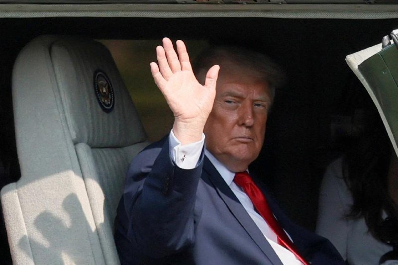 A man in a dark suit and red tie raises his hand while seated inside a vehicle bearing a presidential seal on the headrest.