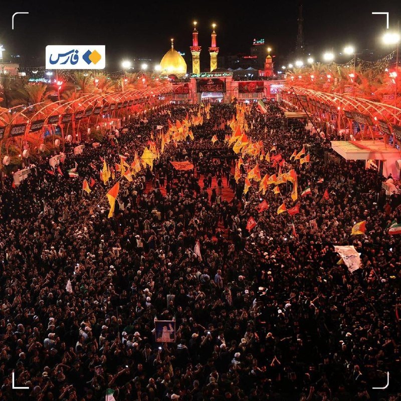 A massive nighttime crowd gathers at a golden-domed shrine, waving yellow and red flags along illuminated walkways between rows of palm trees.