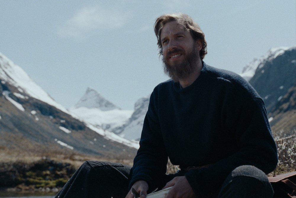 A bearded man in a dark sweater sits smiling against a backdrop of snow-capped mountains under a pale blue sky.