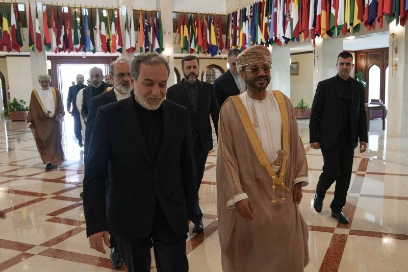 Men in suits and traditional Arab dress walk through a marble-floored hall lined with hanging national flags.