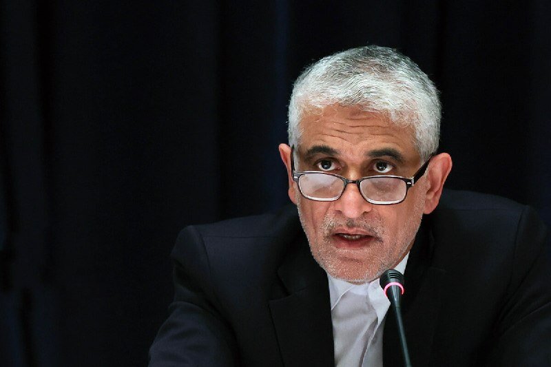 A gray-haired man in a black suit and glasses speaks into a microphone against a dark background.