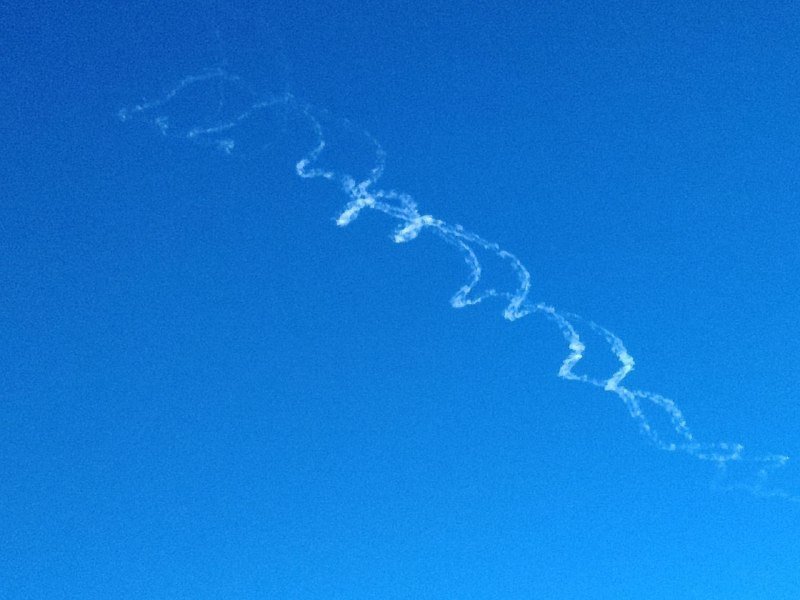 A wispy, squiggly white contrail stretches diagonally across a clear, deep blue sky, breaking apart into scattered curls and fragmented streaks as it dissipates.