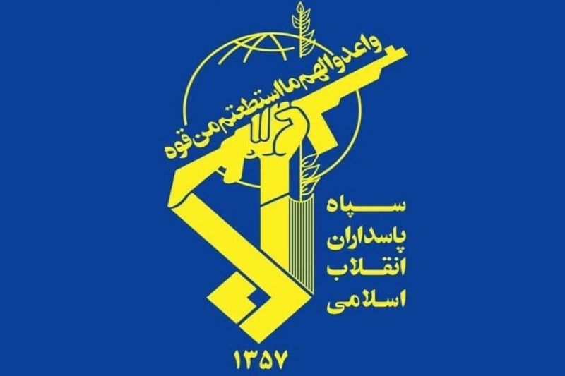 A man in a blue suit and red tie stands on stage with arms outstretched in front of a large American flag and a crowd holding campaign signs.