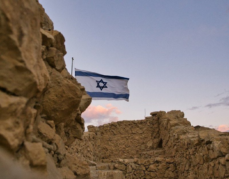 The Israeli flag waves above weathered stone ruins at dusk, with a clear sky and scattered pink clouds in the background.