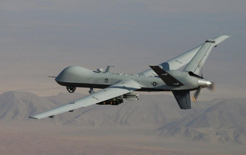 A gray military drone with a propeller flies through the sky over a mountainous, arid landscape, carrying munitions under its wing.