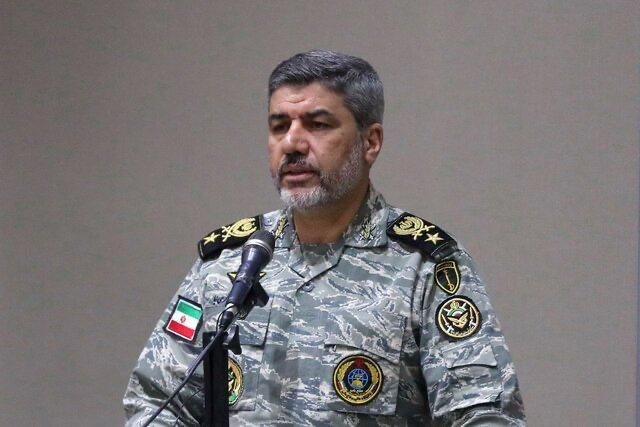 A bearded man in a camouflage military uniform with insignia stands at a microphone against a plain wall, with an Iranian flag patch visible on his sleeve.