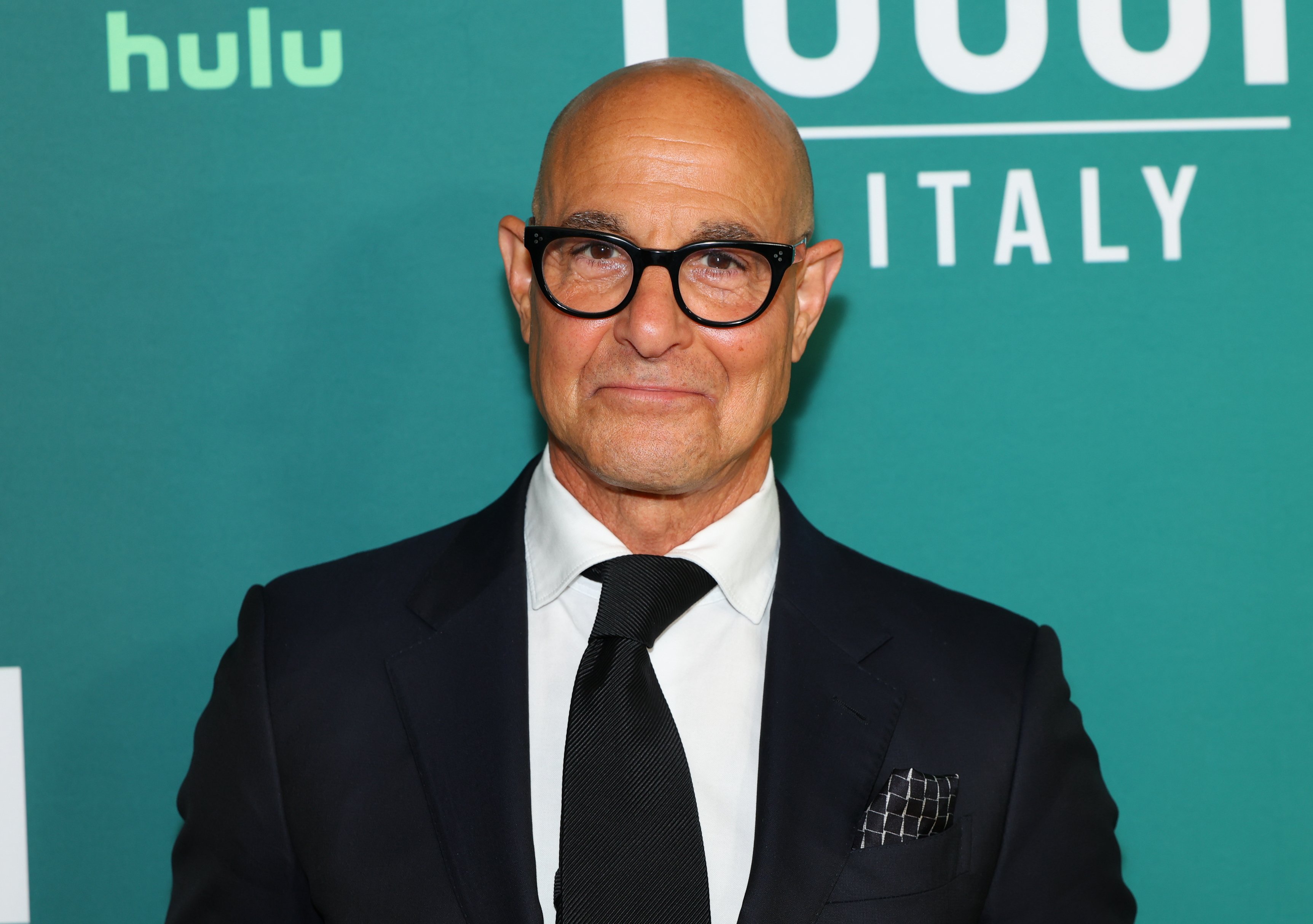 A bald man in a dark suit, white shirt, and black tie poses in front of a green backdrop featuring "hulu" and partial text reading "DULU" and "ITALY."