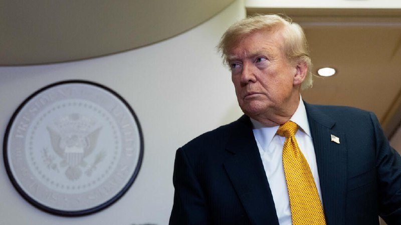 A blonde man in a dark suit and yellow tie stands beside a wall displaying the Presidential Seal of the United States.