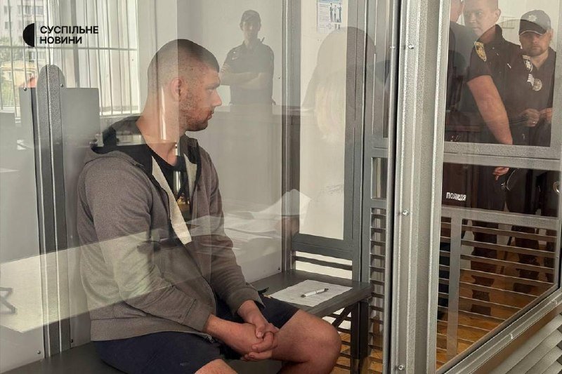 A man in a hooded jacket sits behind a glass enclosure, flanked by uniformed officers standing outside, with "СУСПІЛЬНЕ НОВИНИ" logo visible.