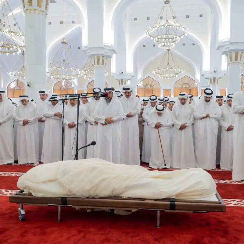 Rows of men in white traditional robes stand behind a body covered in white cloth on a wheeled bier inside a grand mosque with chandeliers and arched pillars.