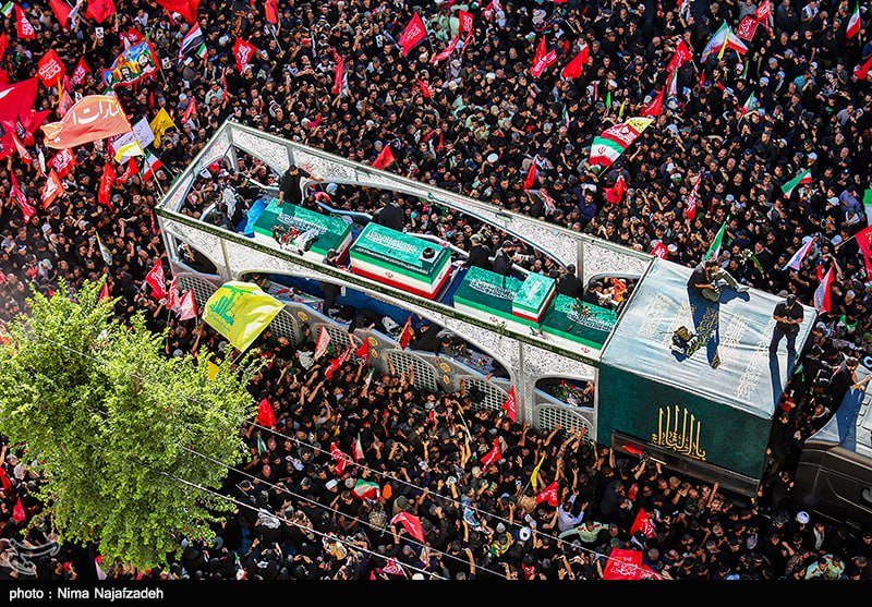 A woman in a black headscarf and robes holds a bouquet of red roses and a portrait of a bearded cleric in a turban amid a crowd of similarly dressed mourners.