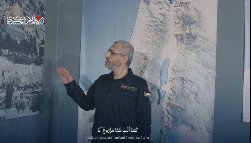 A man in a dark uniform with a Palestinian flag patch gestures toward a wall display of maps, with Arabic and English subtitles reading "just as you are rooted here, so I am."