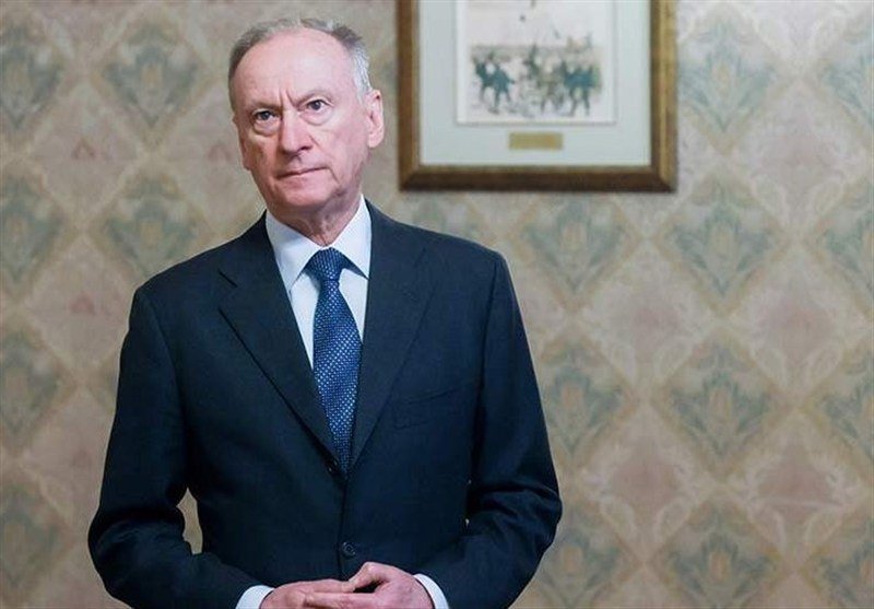 A man in a dark suit and blue patterned tie stands in front of a floral wallpapered wall beneath a framed picture.