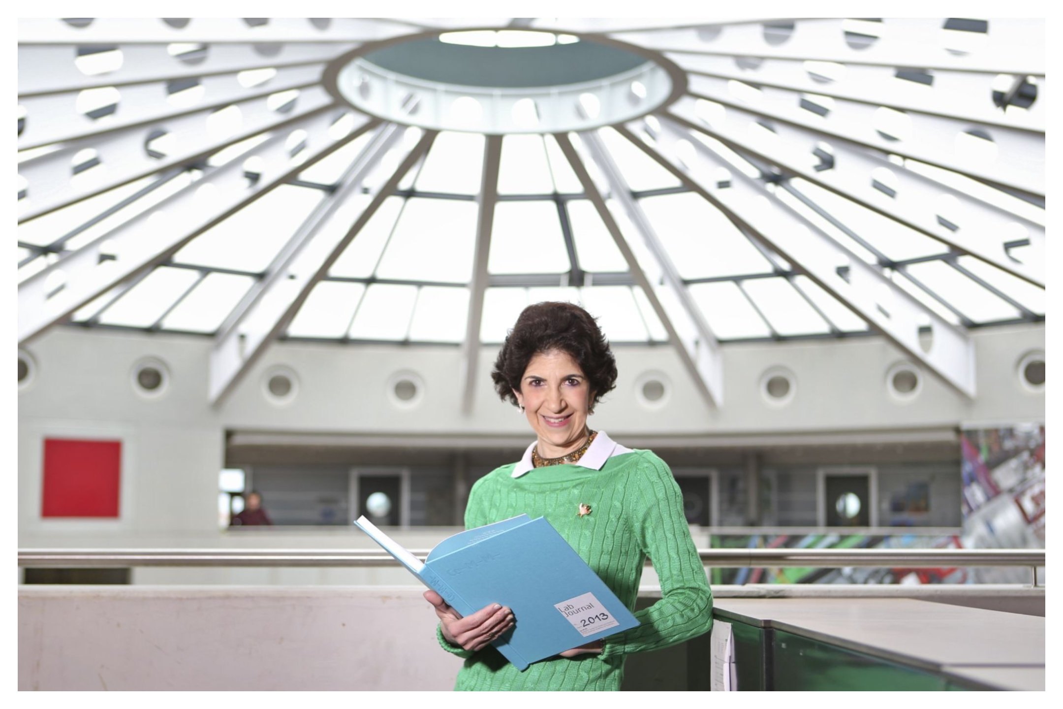 A woman in a green sweater smiles while holding an open blue "Lab Journal 2013" binder beneath a large glass dome ceiling.