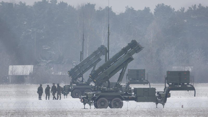 Soldiers stand near large green missile launcher vehicles deployed in a snowy field with barbed wire and a forested background.