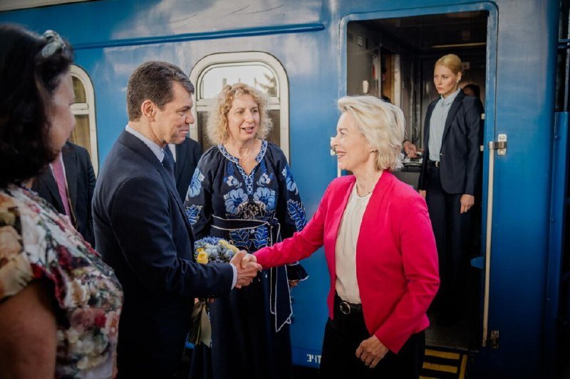 A woman in a pink blazer shakes hands with a man holding flowers beside a blue train, while other formally dressed individuals stand nearby.