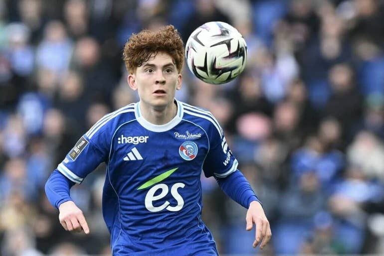 A young soccer player in a blue jersey tracks a ball in mid-air during a match, with a blurred crowd in the background.