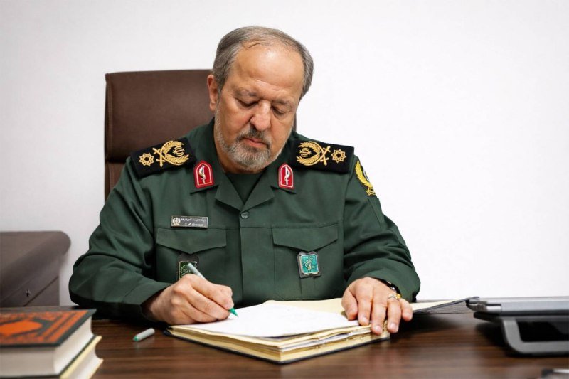 A gray-bearded man in a green military uniform with gold and red insignia writes on papers at a wooden desk.