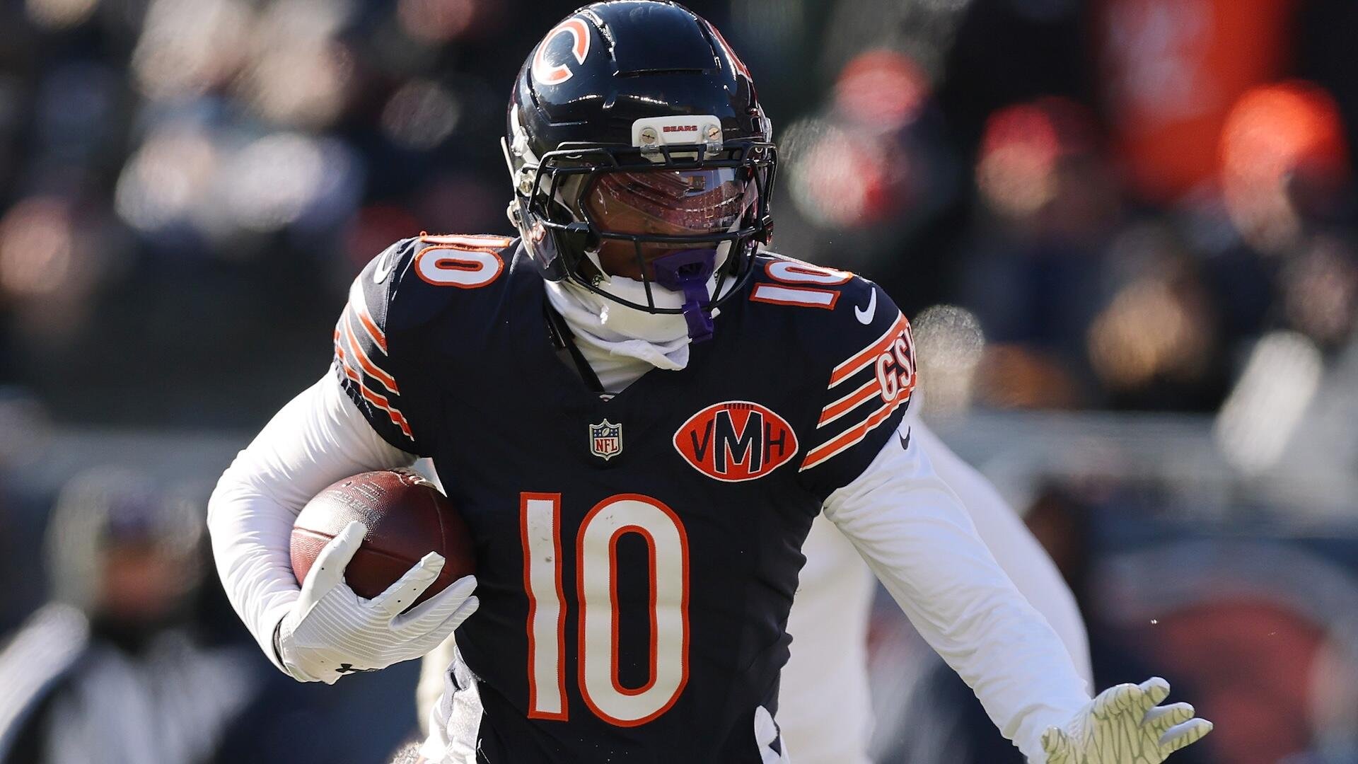 A football player wearing a navy Chicago Bears jersey with the number 10 and a navy helmet runs while carrying a football, with blurred spectators visible in the background.