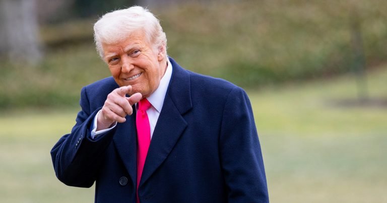 A smiling man with white hair in a navy coat and red tie points toward the camera on a blurred grassy background.
