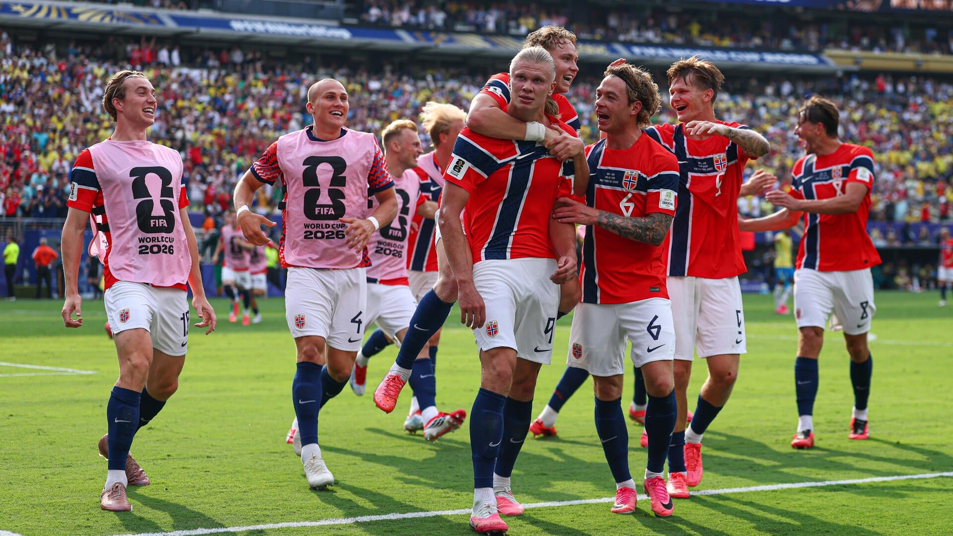 Soccer players in red, white, and blue uniforms celebrating on a stadium field, some wearing pink "FIFA World Cup 2026" bibs, before a large crowd.