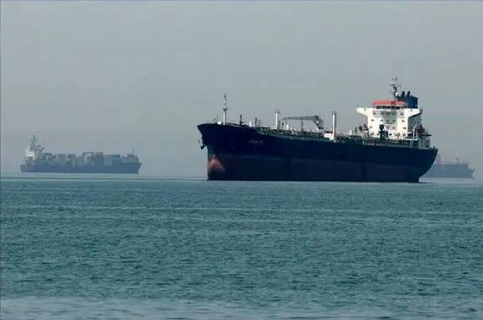 A large cargo ship with a white superstructure sails across hazy gray waters, with a container ship visible in the distant background.