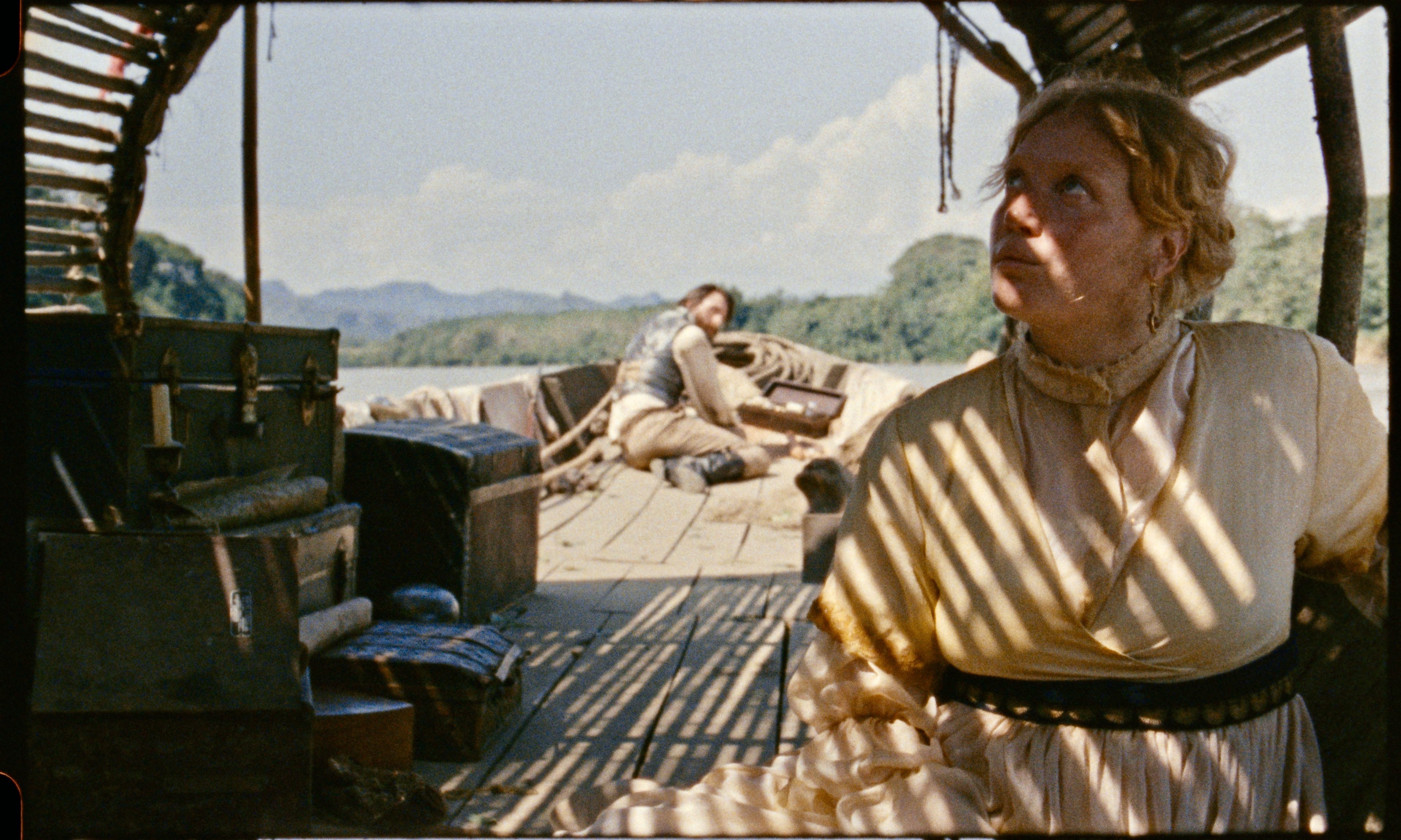 A woman in a cream-colored dress stands on a wooden boat covered in trunks and supplies, with a man resting on deck and a river and forested hills visible in the background.