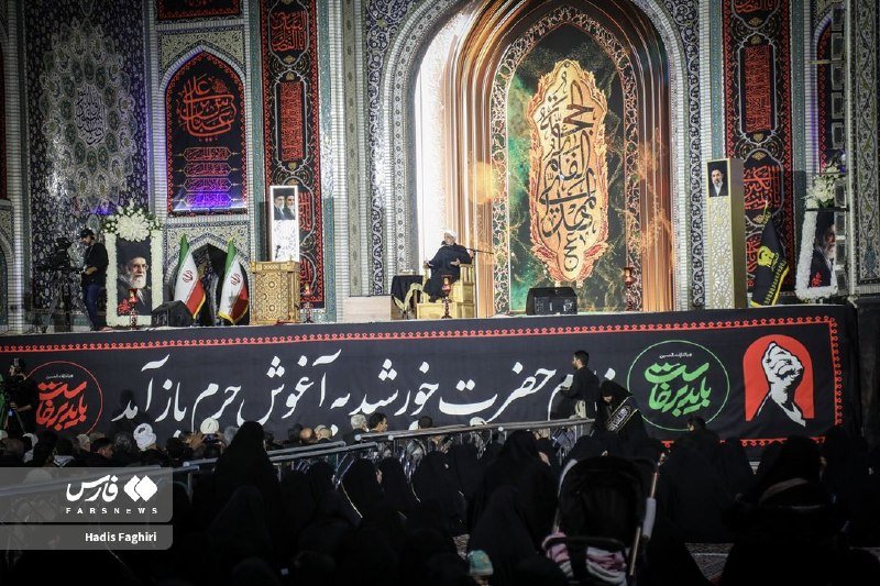 A cleric speaks at a podium on an ornate stage decorated with Persian calligraphy and Iranian flags, addressing a seated crowd of women in black chadors.